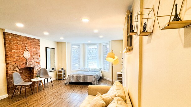 A cozy room with a brick fireplace, a bed with a striped bedspread, a sofa, and a small table with chairs. - Charming Brick Row House close to NYC (Bayonne)