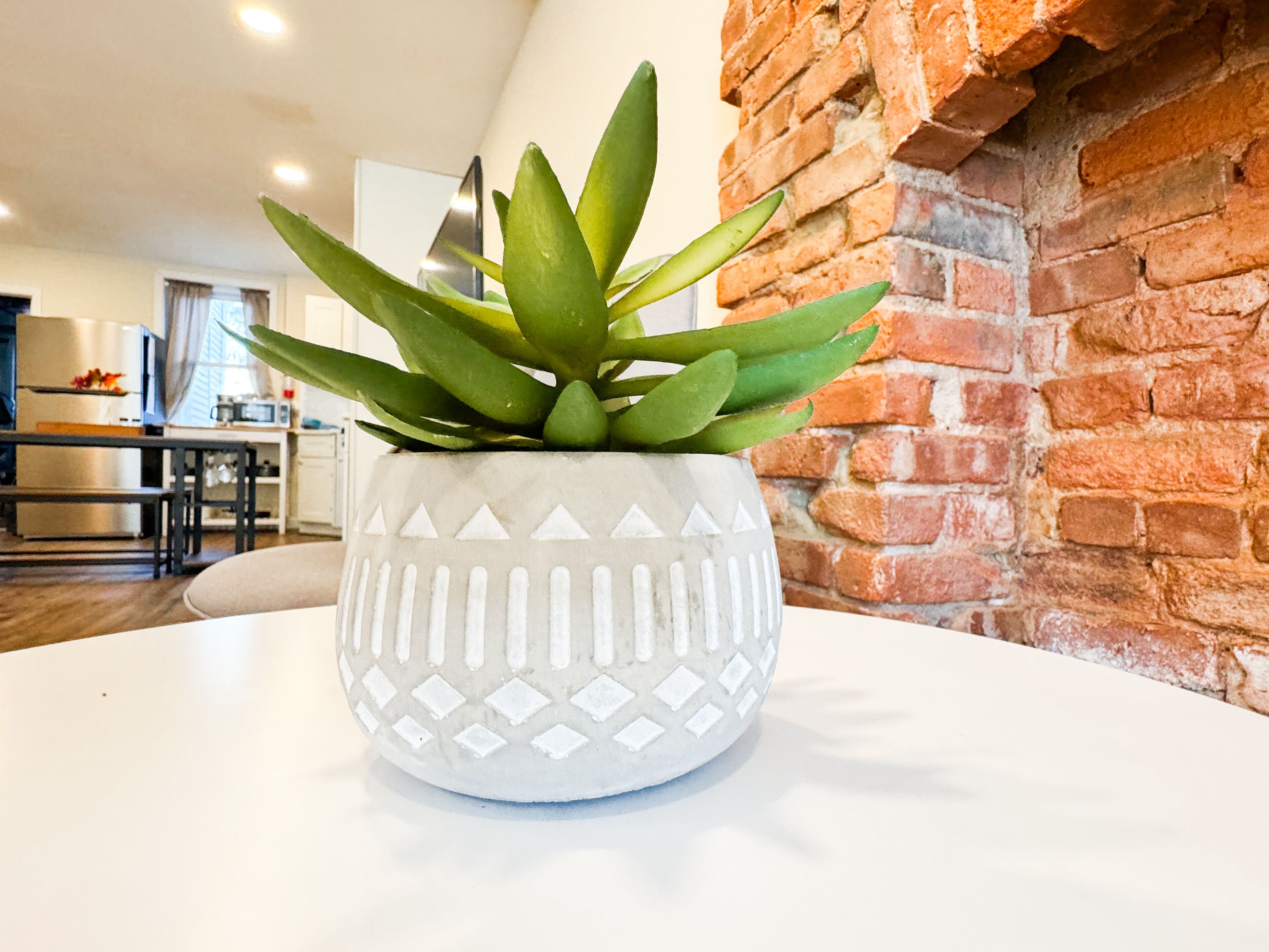 Une plante en pot est posée sur une table blanche dans une cuisine moderne, avec un mur en briques et des appareils électroménagers en acier inoxydable.