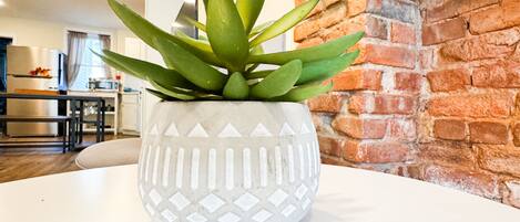 Une plante en pot est posée sur une table blanche dans une cuisine moderne, avec un mur en briques et des appareils électroménagers en acier inoxydable.