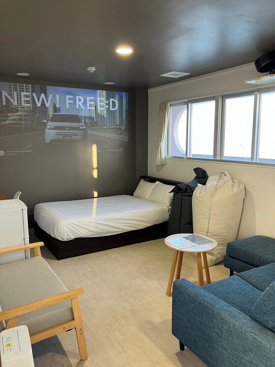 Moderni hotellihuone, jossa on sänky, pieni pöytä, sohva, projektorikangas, jolla näkyy kuva autosta, ja ikkuna.