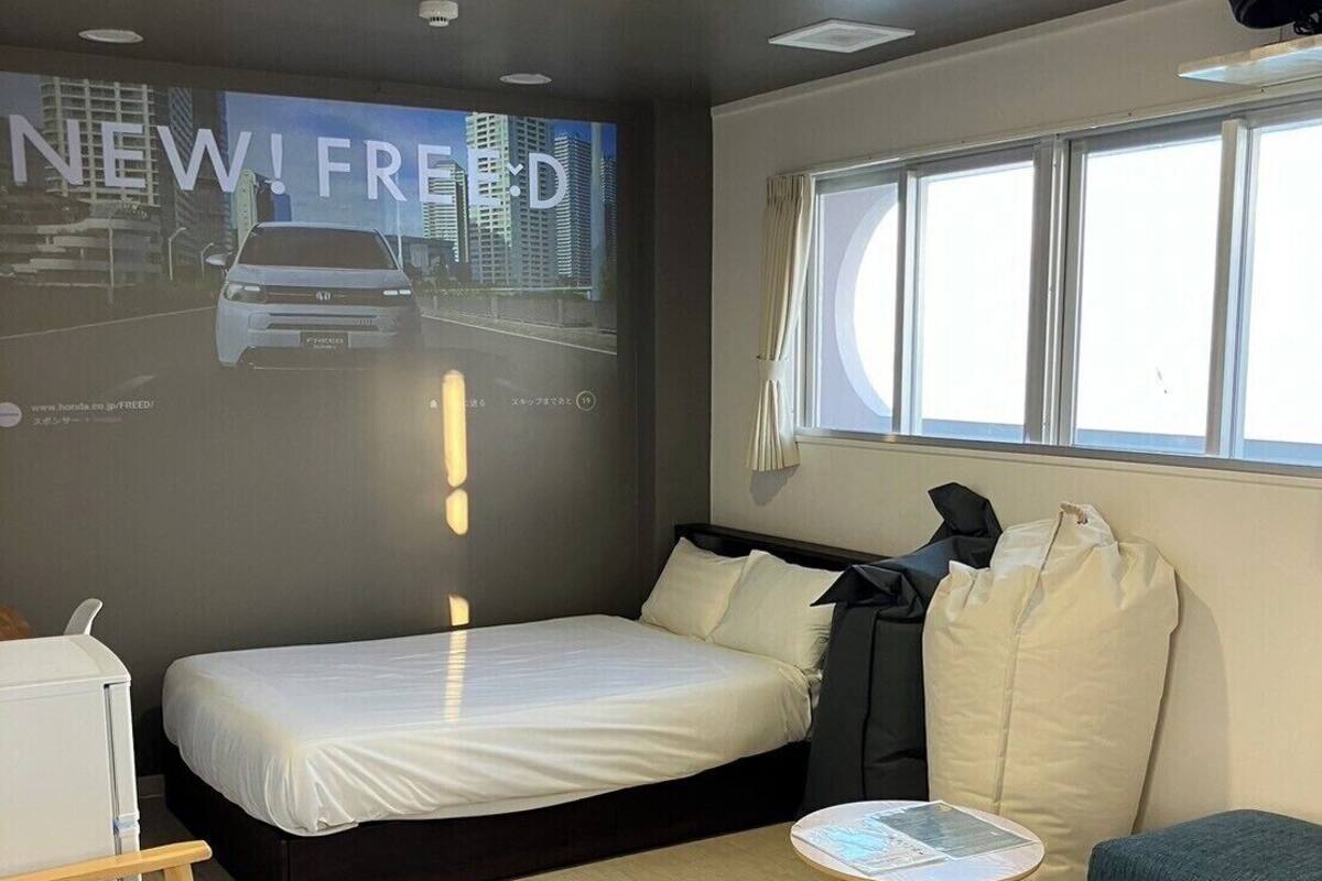 Ein modernes Hotelzimmer mit Bett, einem kleinen Tisch, einer Couch, einer Projektionsleinwand, auf der ein Auto zu sehen ist, und einem Fenster.