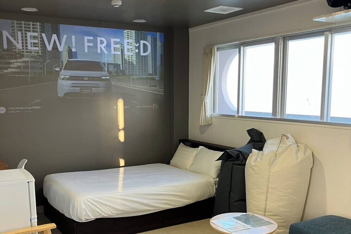 Moderni hotellihuone, jossa on sänky, pieni pöytä, sohva, projektorikangas, jolla näkyy kuva autosta, ja ikkuna.