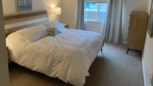 A neatly arranged bedroom with a large bed, a bedside lamp, a painting on the wall, and a view through a window. - Beach House, 1 block to beach, walk to town, great outdoor space (Cayucos)