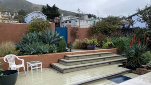 A patio with steps leading to a seating area, surrounded by various plants and a view of residential buildings. - Beach House, 1 block to beach, walk to town, great outdoor space (Cayucos)