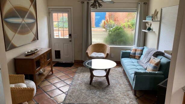 A compact living space with a sofa, a wicker chair, a glass table, and a door leading to a balcony with plants. - Beach House, 1 block to beach, walk to town, great outdoor space (Cayucos)