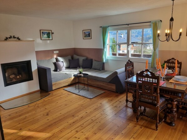 A cozy living room with a built-in sofa, a fireplace, a dining table with chairs, and a large window with curtains. - Fewo Alpenglück und Heimatgfui (Schönau am Königssee)