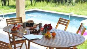 A poolside table with fruits, pastries, and drinks, surrounded by chairs, with a pool in the background.
