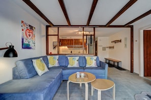 Un salon avec un canapé bleu, deux petites tables, une cuisine à l’arrière-plan, des poutres en bois au plafond et un tableau coloré au mur.