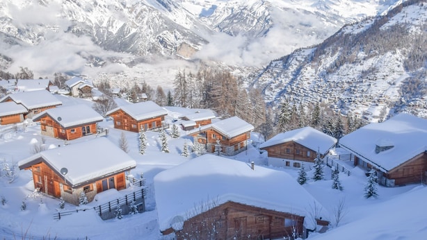 Uma aldeia montanhosa coberta de neve, com cabanas de madeira, rodeada por montanhas e árvores cobertas de neve.