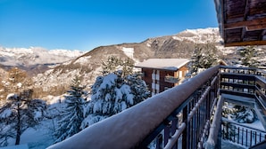 Uma cabana de montanha com fachada de madeira, uma varanda com guarda-corpo de metal e uma paisagem nevada com montanhas ao fundo.