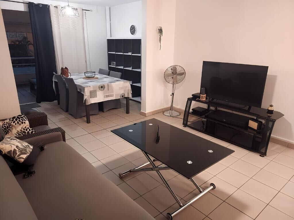 A living room with a sofa, a coffee table, a TV, a fan, and a dining area with a table and chairs.