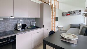 Et moderne køkken med indbygget ovn, kaffemaskine og vask. Der er et spisebord med en tallerken og en stol. En stige står lænet mod væggen, og en sofa ses i baggrunden.