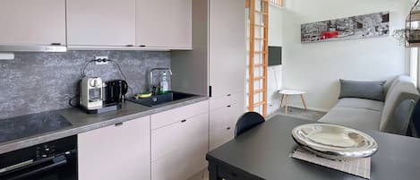 A modern kitchen with a built-in oven, a coffee maker, and a sink. There is a dining table with a plate and a chair. A ladder is leaning against the wall, and a sofa is in the background.