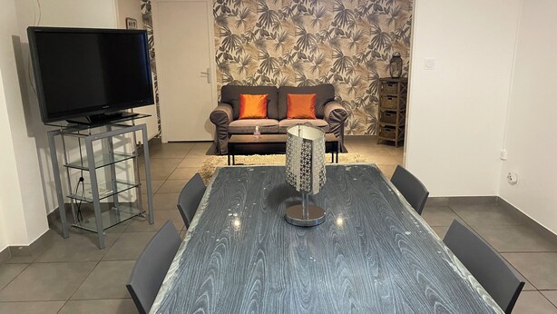 A dining area with a glass-top table, chairs, a television on a stand, a sofa with cushions, and a patterned wallpaper. - Le Mathiez (Dijon)