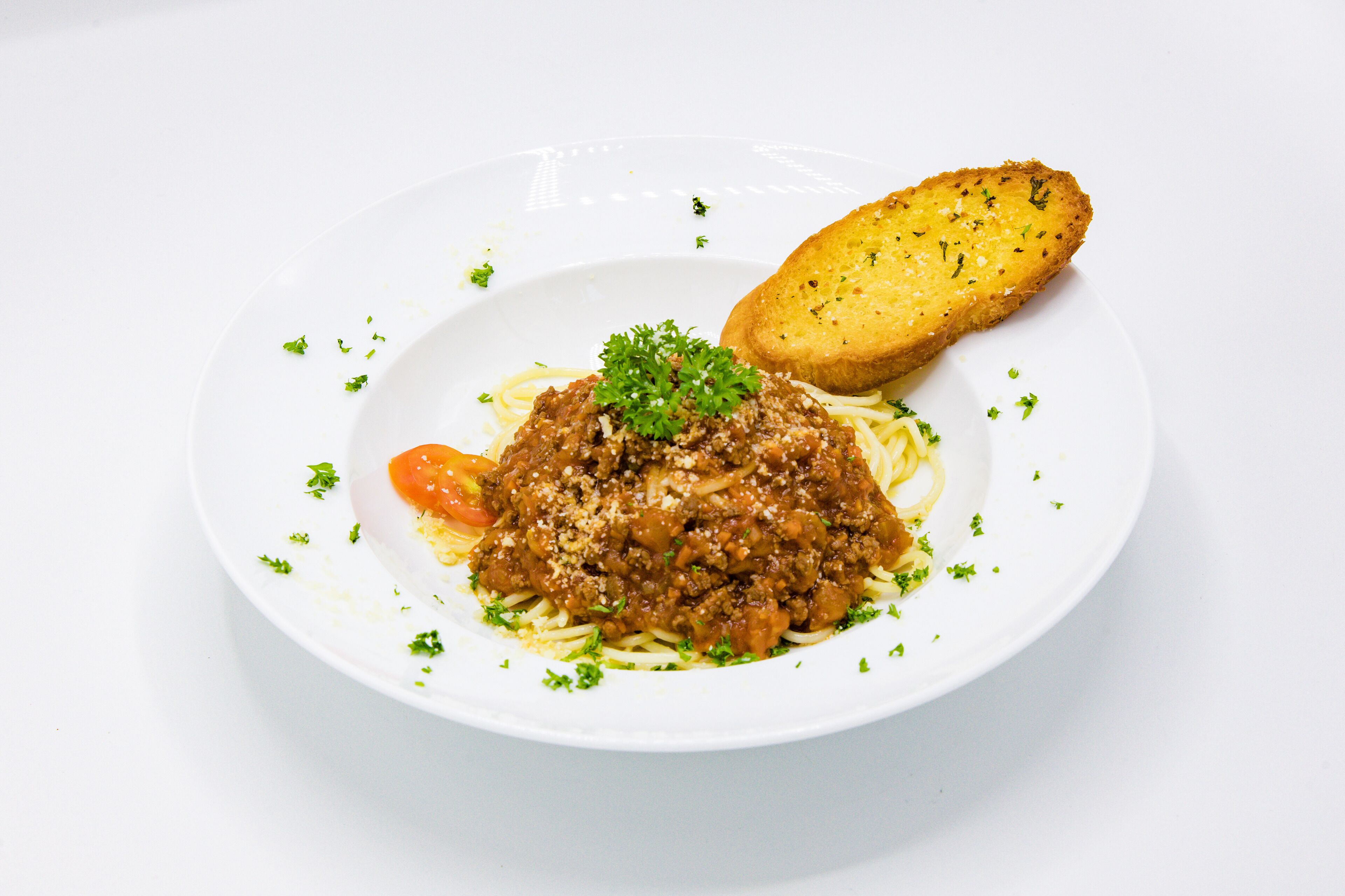 Lautasella on spagettia lihakastikkeella, päälle ripoteltu persiljaa, ja valkoisella lautasella on myös viipale leipää.