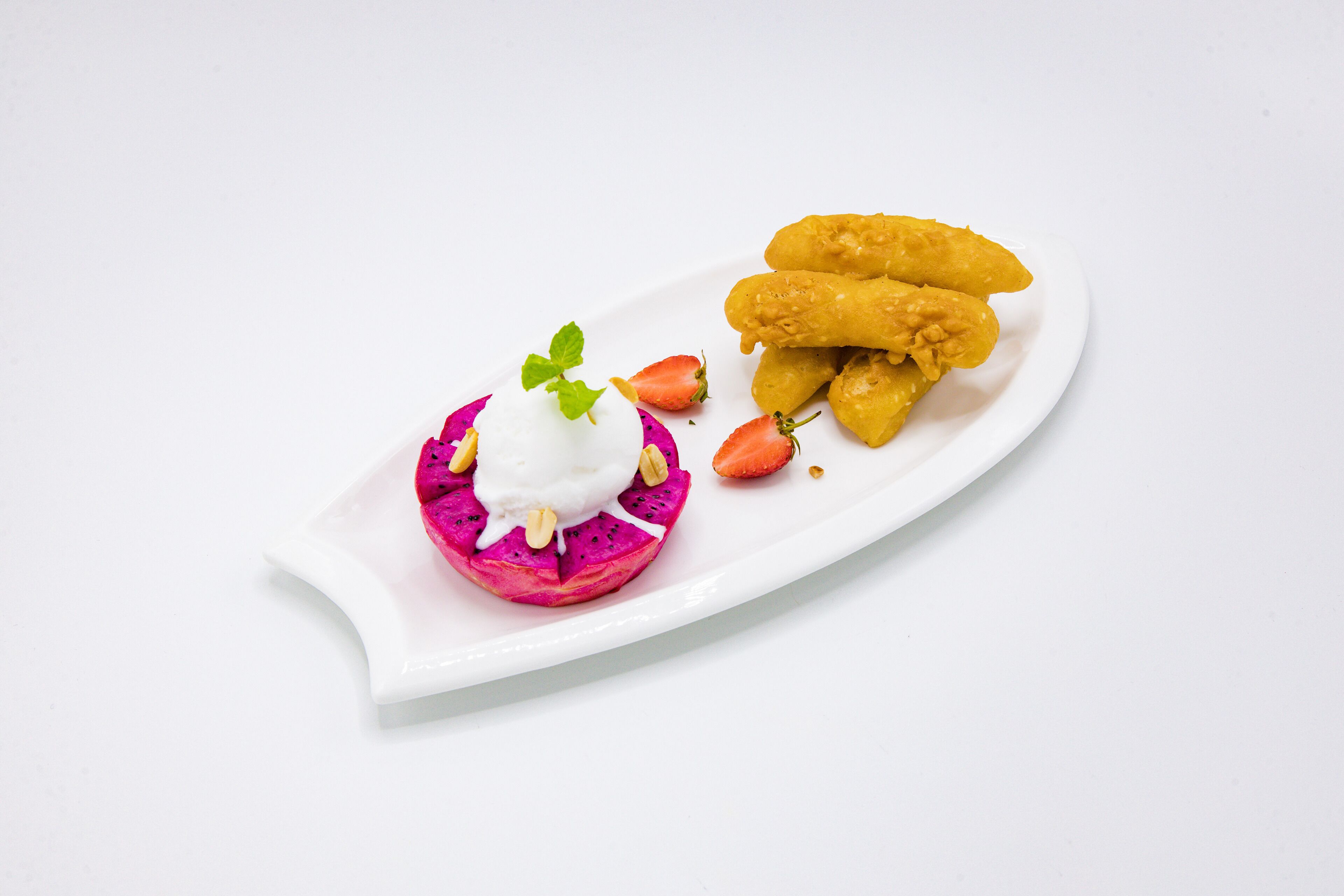 Une assiette blanche contenant un dessert composé d’une boule de fruit du dragon, surmontée de crème fouettée et d’une feuille de menthe, accompagnée de trois morceaux de nourriture frits, dorés, et de quelques fraises.