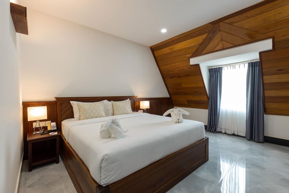 Una habitación de hotel moderna con techo de madera, una cama grande con ropa de cama blanca, dos mesitas de noche con lámparas y una ventana con cortinas.