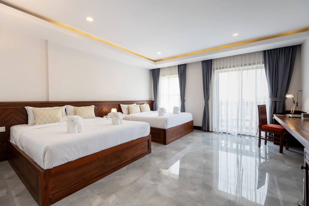 Habitación de hotel con dos camas, cabecero de madera, escritorio con silla y ventanas grandes con cortinas.