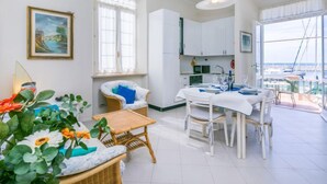 Un intérieur lumineux et blanc comprenant un coin repas, une cuisine et de grandes fenêtres offrant une vue sur la mer.