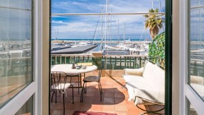 Un balcon avec un canapé blanc, une petite table et des chaises, donnant sur une marina où sont amarrés des bateaux, avec un ciel dégagé.