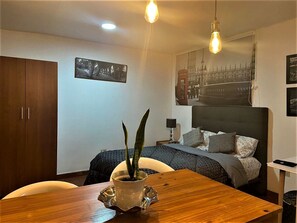 A hotel room with a bed, a wooden table with a plant, a wardrobe, and two wall-mounted light fixtures. - Mercanti Apts Caney (Cali)