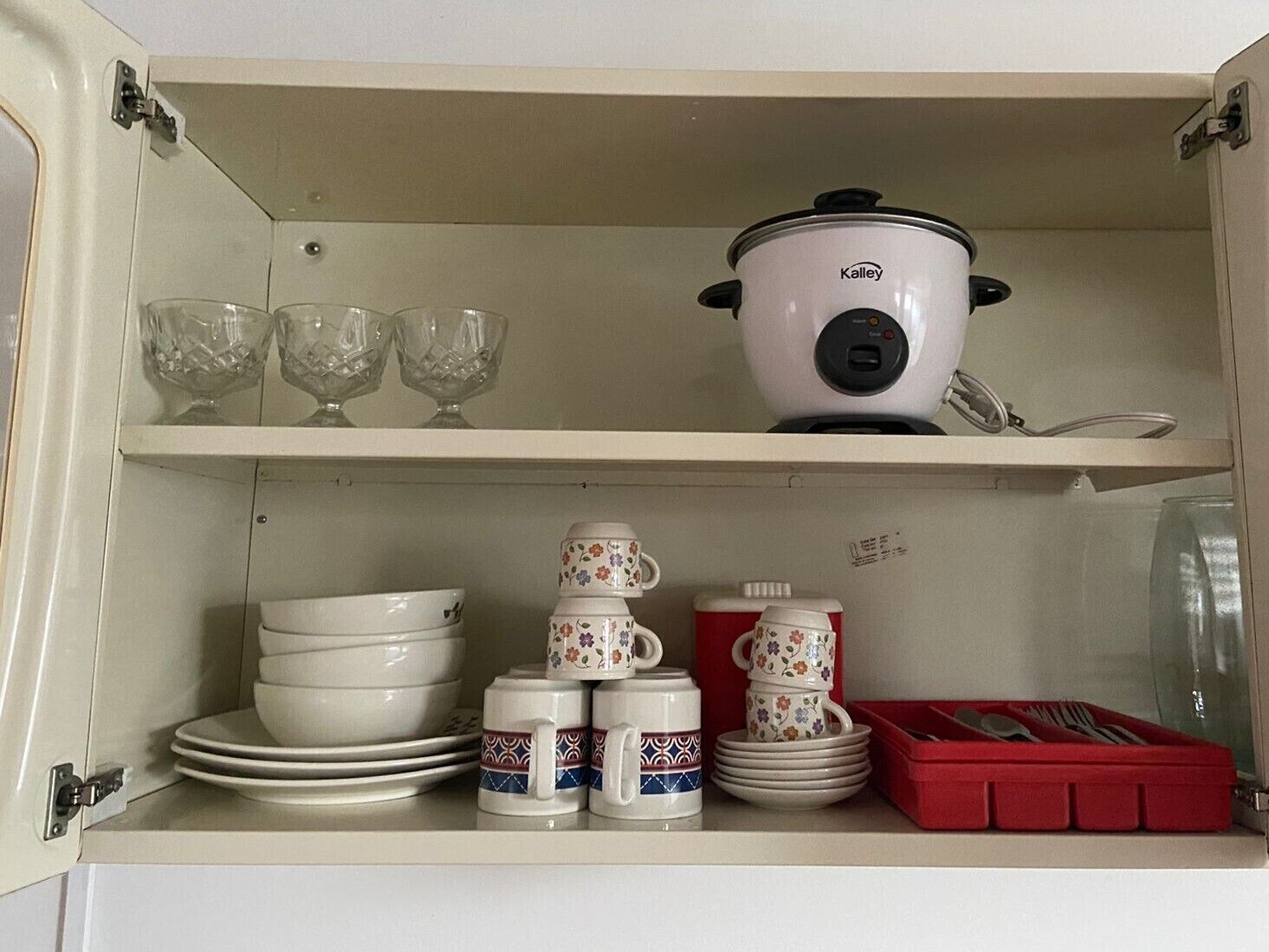 A kitchen cabinet with a white pot, stacked plates, cups, and a red utensil box.