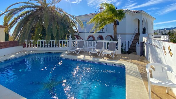 A swimming pool with white lounge chairs, a white building with arches, and palm trees.