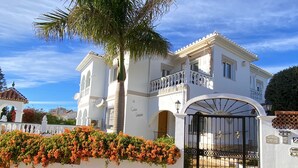 A large, white, two-story building with a gated entrance, a balcony, and a palm tree in front.
