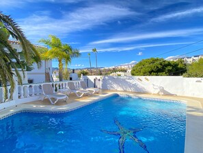 Une piscine entourée de chaises longues, de palmiers et surplombée d’un ciel bleu azur.