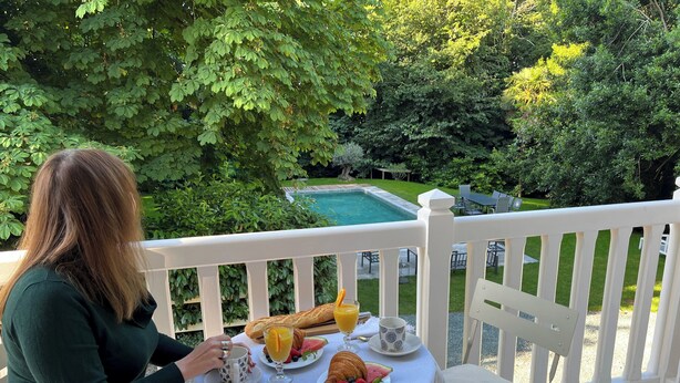 Iemand zit aan een ronde tafel met een wit tafelkleed en geniet van een maaltijd bestaande uit croissants, fruit en sap. De tafel staat op een terras met een witte balustrade, met uitzicht op een zwembad en weelderige groene beplanting.