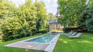 Een rechthoekig zwembad met houten terras, omringd door een verzorgd gazon en bomen.