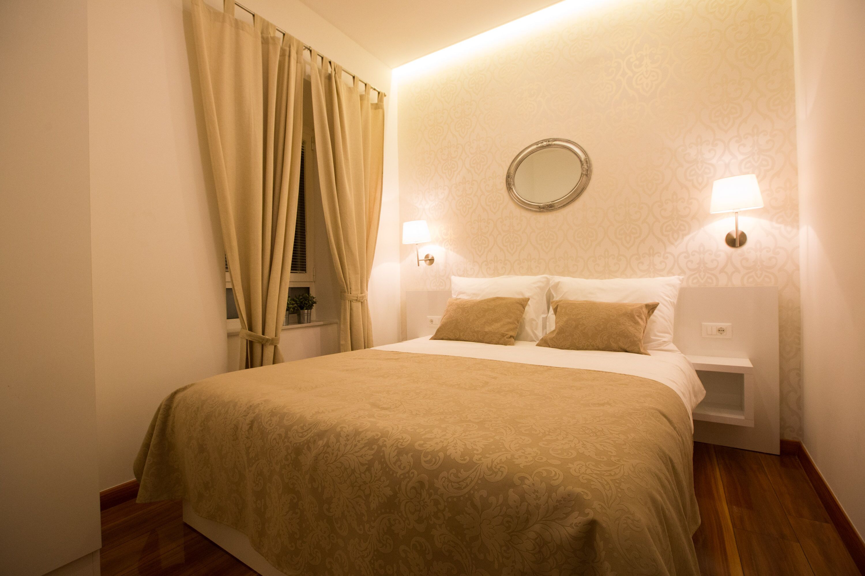 A hotel room with a large bed, two bedside lamps, a round mirror on the wall, and a window with curtains.