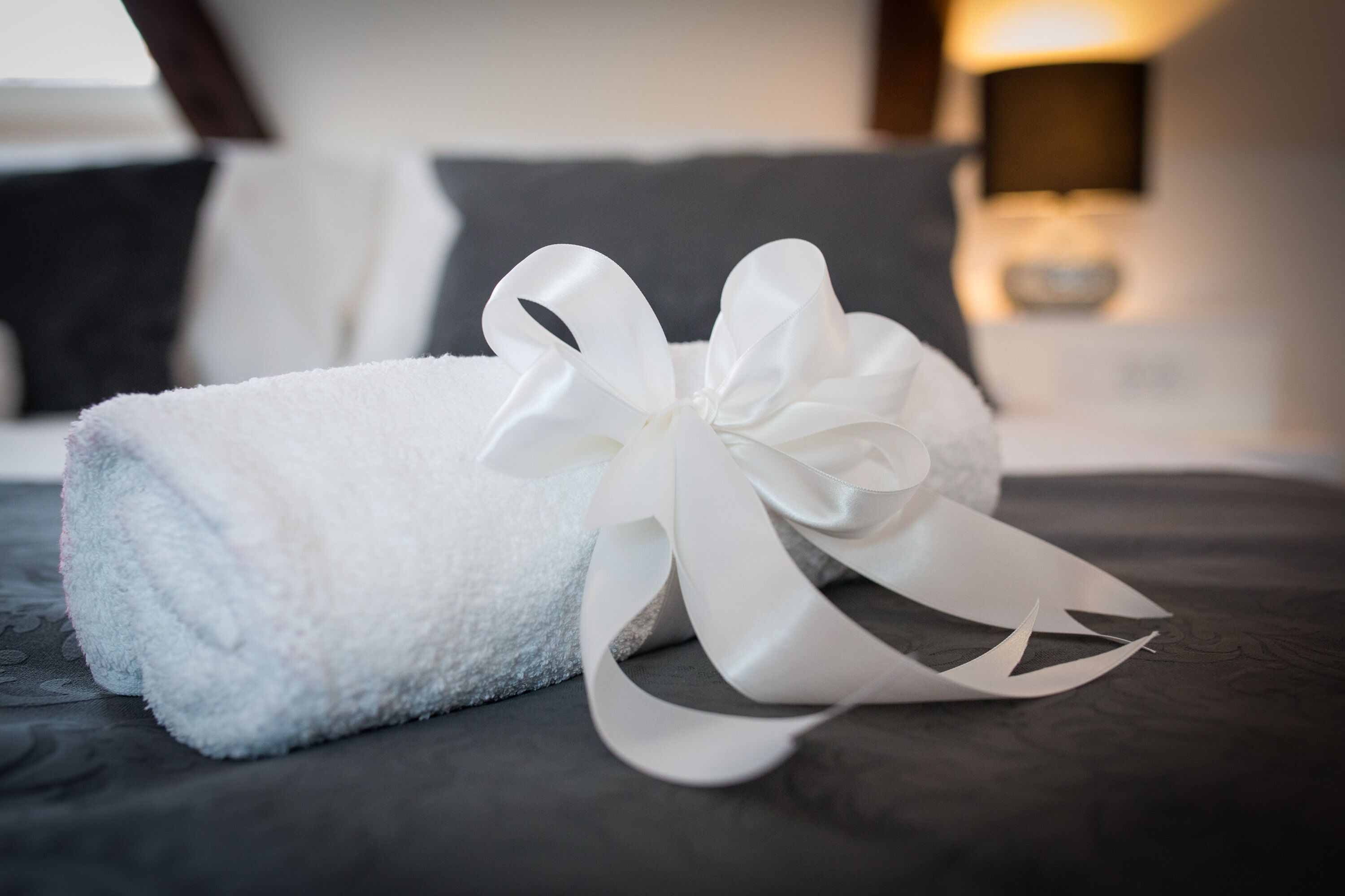 A neatly folded white towel with a large bow on a dark-colored bed.