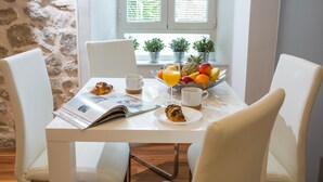 A breakfast table with croissants, fruit, and a magazine. There are two white chairs, a window with plants, and a stone wall. - Casa Arancia (Dubrovnik)