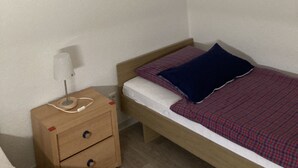 Een kleine eenpersoonskamer met een houten hoofdbord, een nachtkastje met een lamp en een lijst met een afbeelding aan de muur.