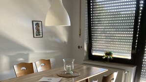 Een eethoek met een houten tafel en stoelen, een witte hanglamp, een lijst met een afbeelding aan de muur, en een raam met jaloezieën.