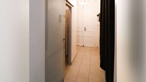 Un couloir étroit, avec une porte au fond, un luminaire au-dessus de la porte et un rideau noir sur la droite.