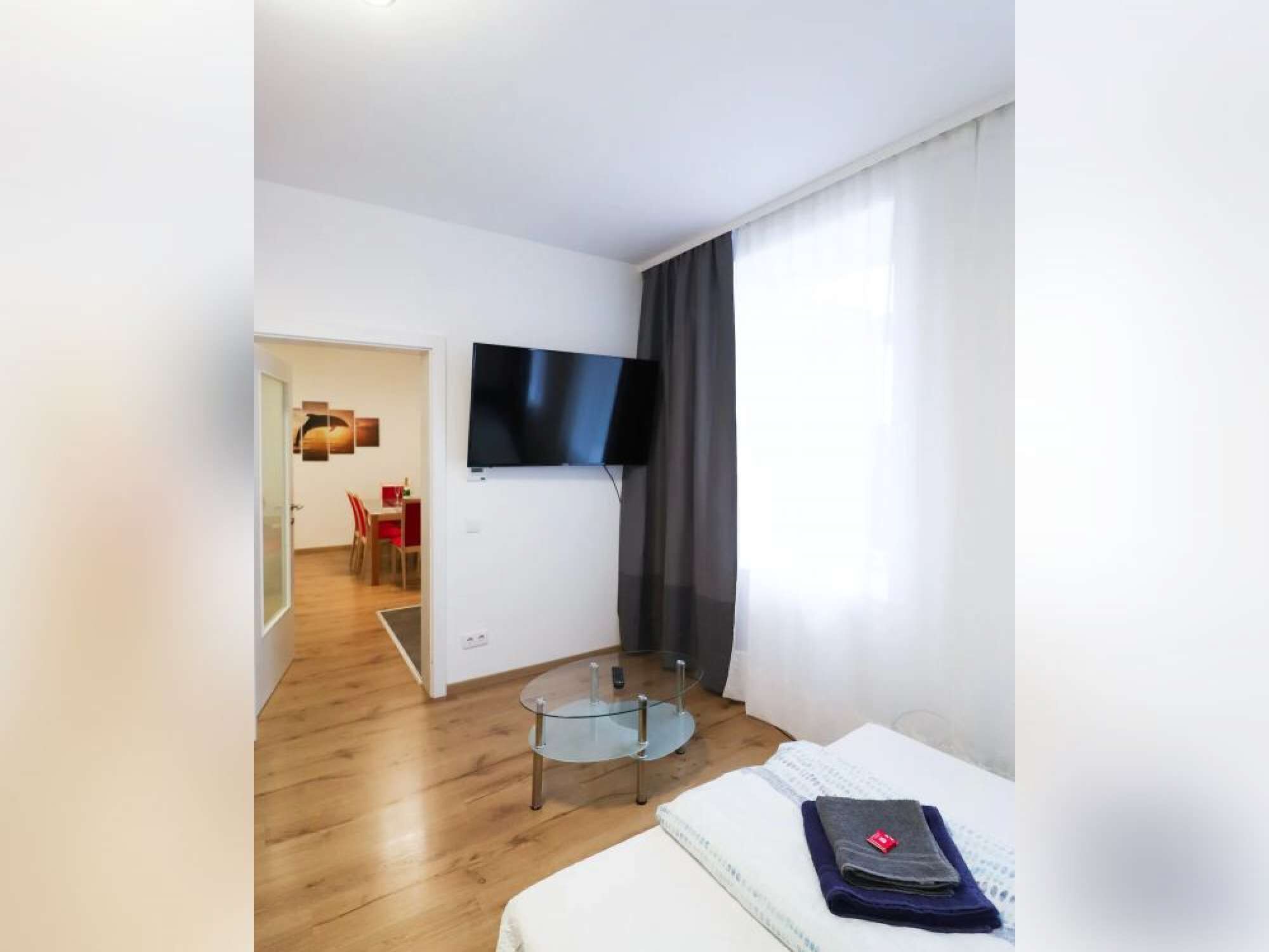 A modern hotel room with a flat-screen TV mounted on the wall, a glass coffee table, a bed with a neatly folded towel, and a dining area visible through an open door.