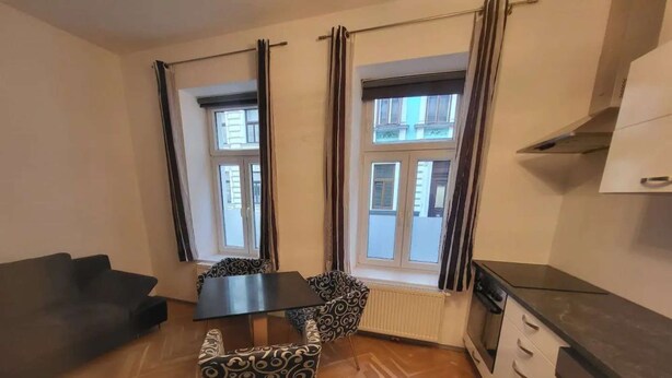 Un espace de vie compact comprenant un canapé, une table à manger avec des chaises à motifs, un coin cuisine avec une cuisinière et des fenêtres avec des rideaux.