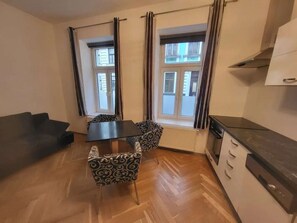 Ein kompakter Wohnbereich mit einer Couch, einem Esstisch mit gemusterten Stühlen, einer Küchenzeile mit Herd und Fenstern mit Vorhängen.