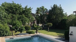 A rectangular swimming pool with a wooden deck on one side and a grassy area with trees and a white wall in the background. - Downtown Toulouse house with pool (Toulouse)