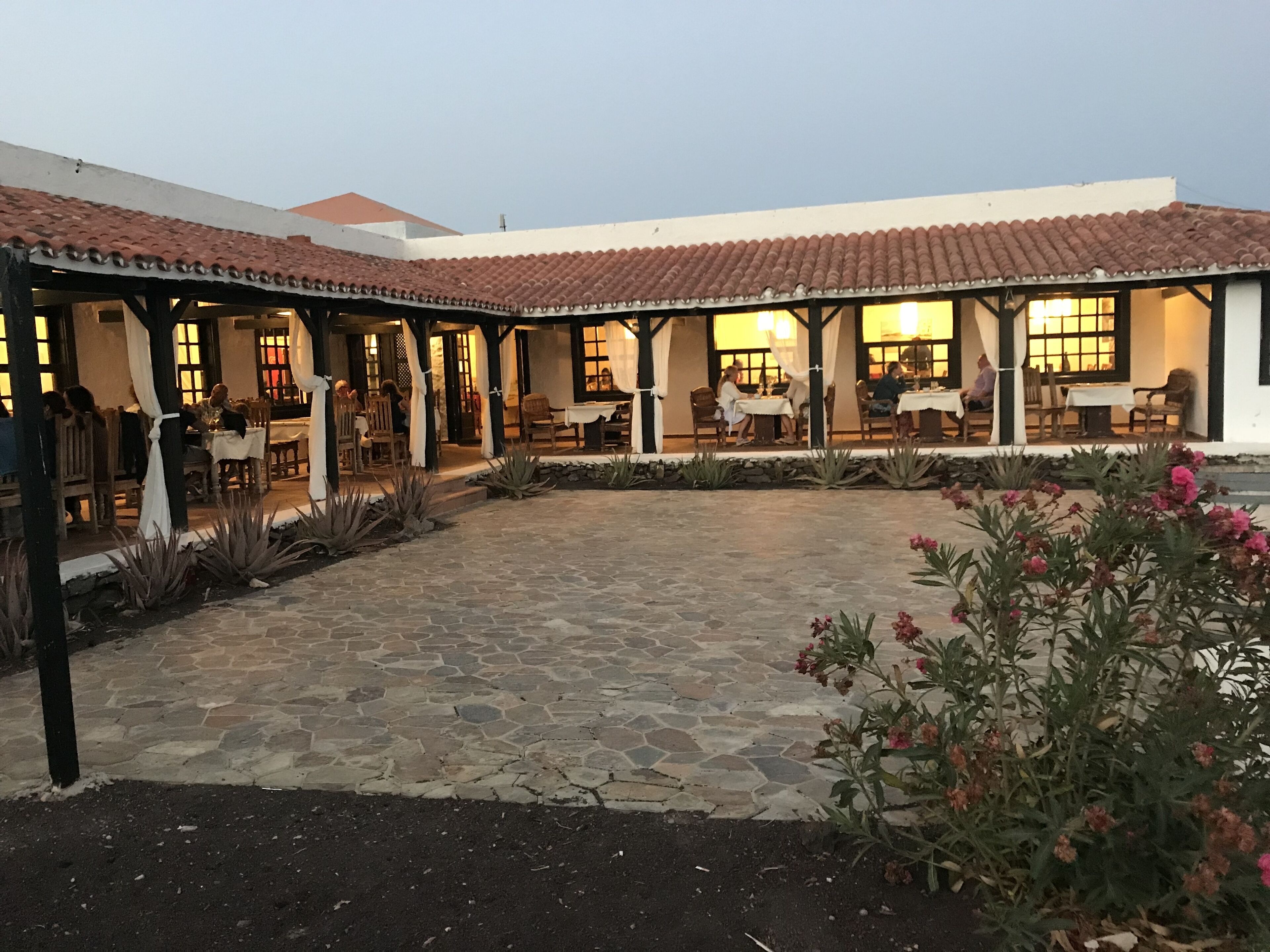 A courtyard with stone flooring, outdoor seating, and a white building with a tiled roof.