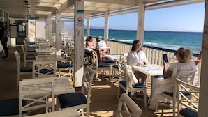 Un espacio de comedor al aire libre con mesas y sillas, vista al mar y un cartel que prohíbe la entrada de perros.