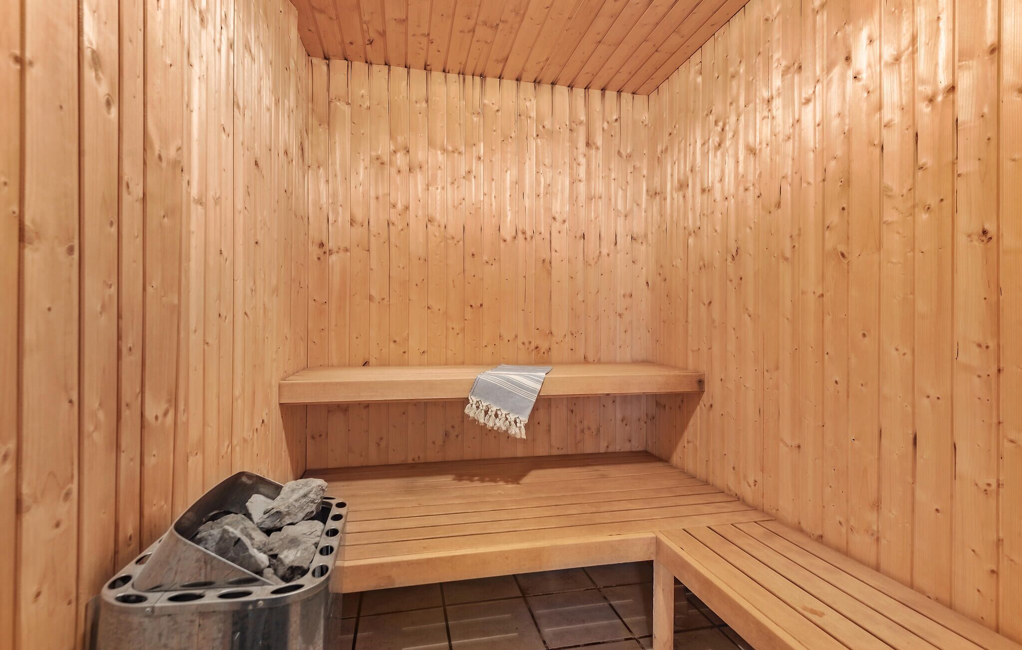 A sauna with wooden walls and benches, a metal ashtray with rocks, and a bench with a striped towel.