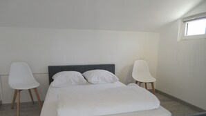 Une chambre à coucher avec un lit, deux chaises blanches, une fenêtre et une tête de lit grise.