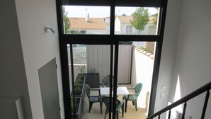 On aperçoit, à travers une porte vitrée, un patio extérieur avec une table et des chaises, une clôture blanche et, au fond, un bâtiment dont le toit est recouvert de tuiles.