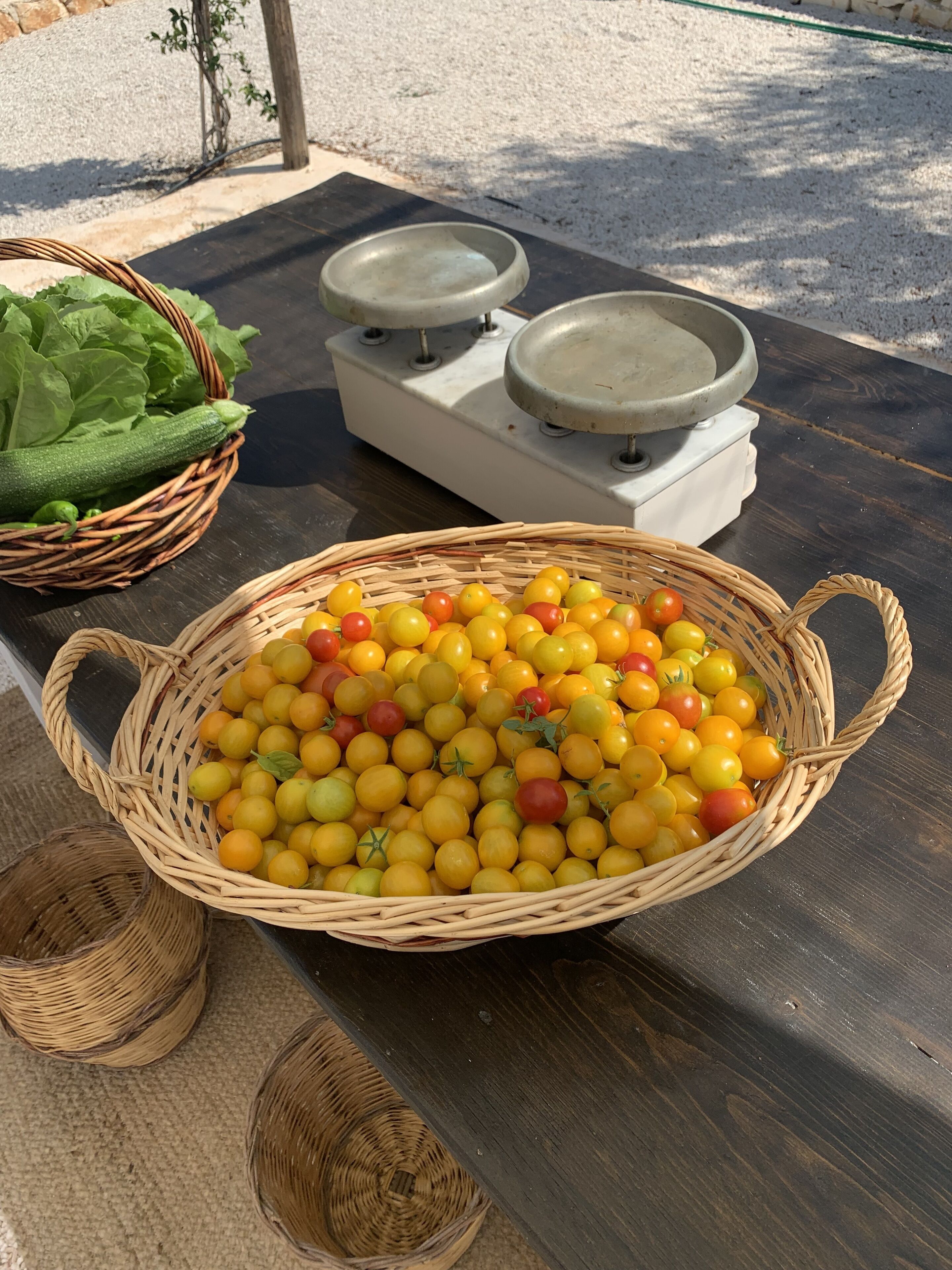 Una mesa con una cesta de tomates cherry amarillos y rojos, una cesta de mimbre con verduras verdes y una estufa blanca con dos ollas de metal.