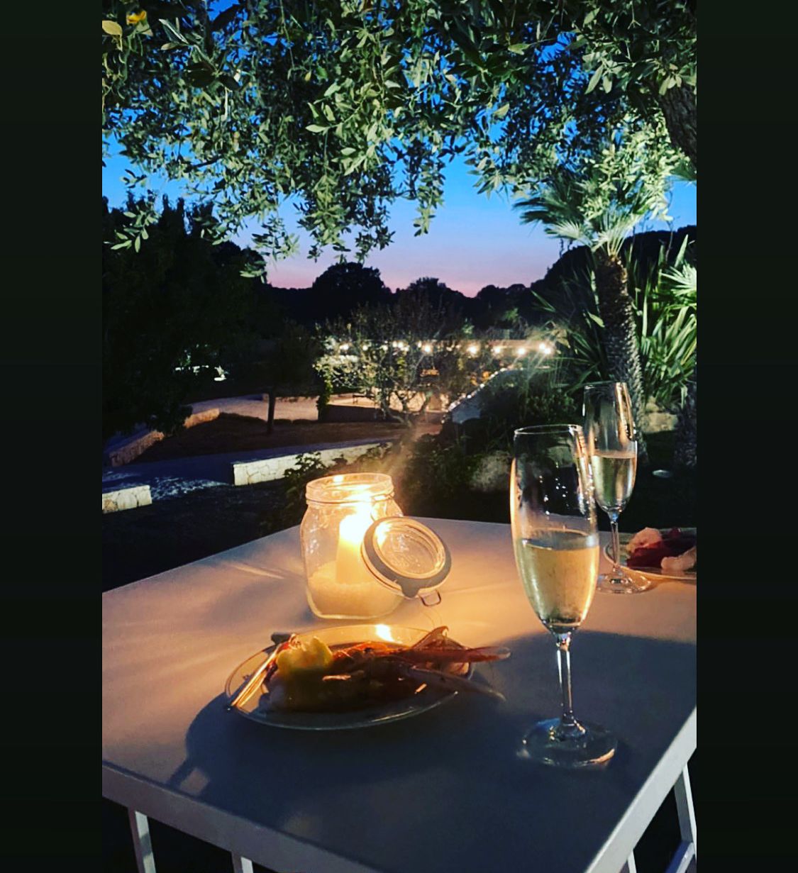Une table à manger en plein air, avec une bougie, un verre de vin blanc et une assiette de nourriture.