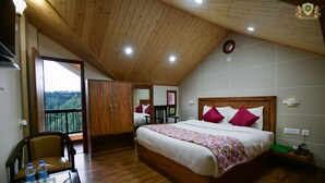 Un dormitorio con techo de madera, una cama con ropa blanca y almohadas rosas, una silla de madera, una mesita con botellas y vista a un bosque a través de una puerta balcón.