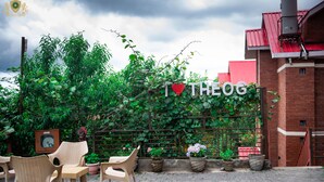 Un área de estar al aire libre con sillas de mimbre y plantas en macetas, un cartel que dice "I ♥ THEOG", y un edificio de ladrillo al fondo.